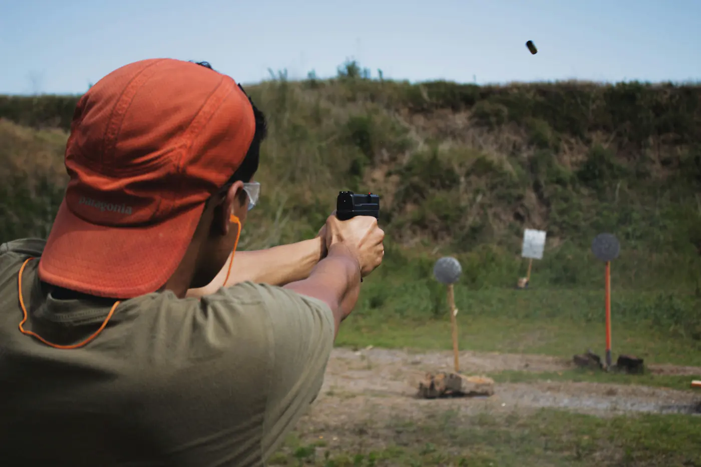 Un homme de dos, casquette orange, tire au pistolet vers des cibles métalliques sur un pas de tir en plein air