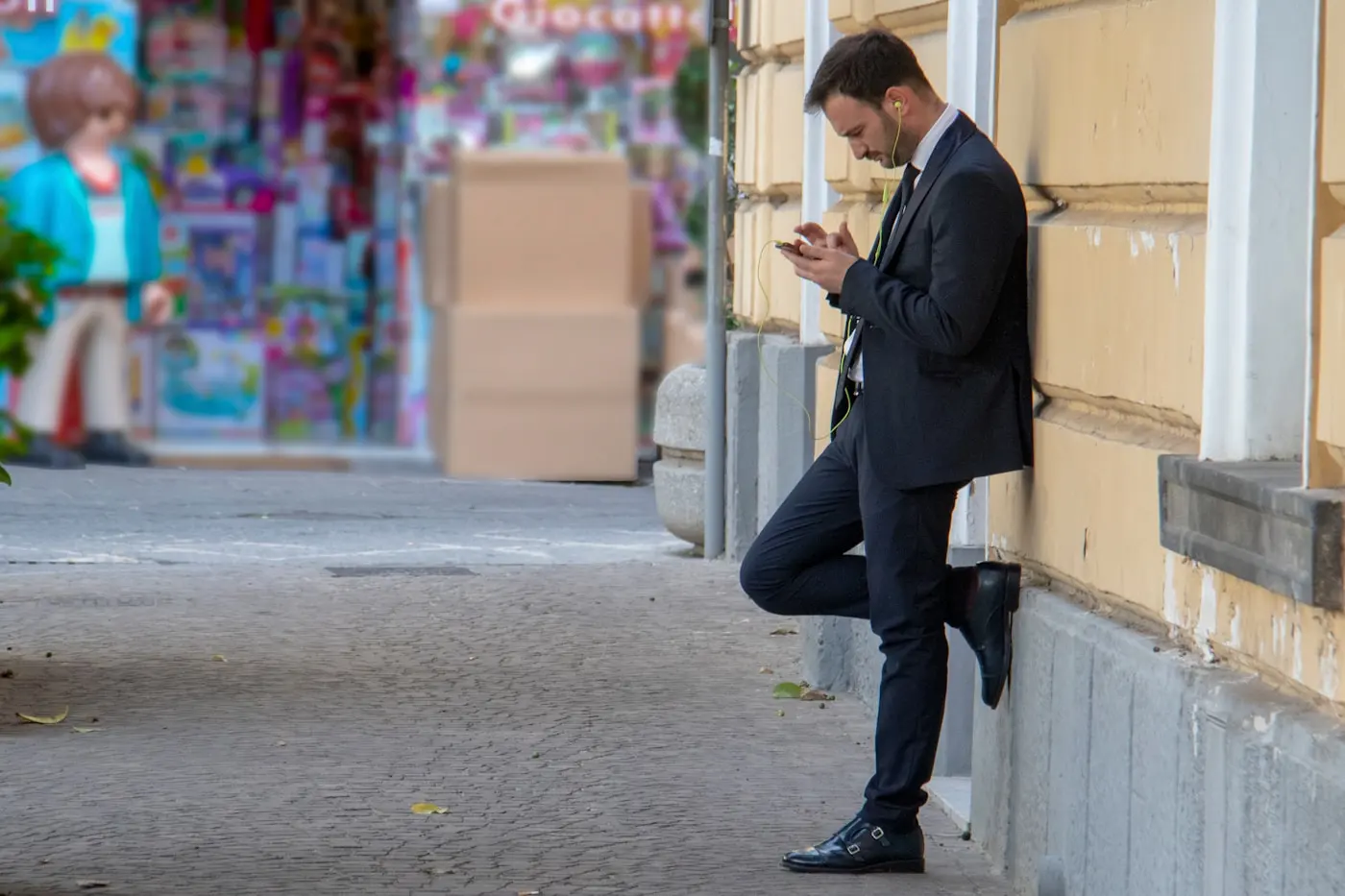 Un homme consulte son téléphone dans la rue, adossé à un mur, devant la vitrine d'un commerce